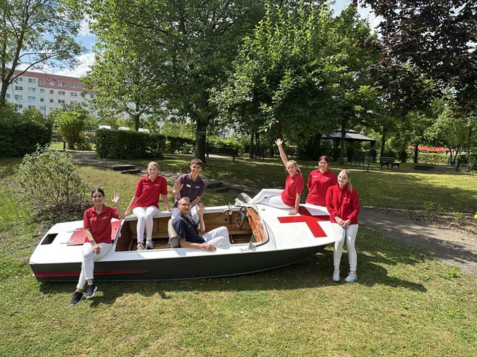 Alle in einem Boot Bild zeigt die 7 Personen in einem Boot sitzend. Dieses Boot steht auf einer Grünfläche im Park des Pflegeheim und signalisiert, dass wir alle in einem Boot sitzen.