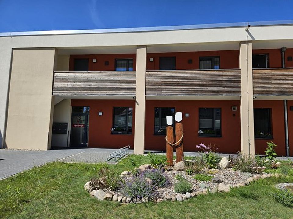 Einrichtung TP Bützow Bild zeigt die Gebäudefrontansicht der Tagespflege. Es ist der Eingang mit dem Laubengang zu sehen. Im Vordergrund ist das mit Blumen und Bodendeckern gestaltete und mit kleineren Feldsteinen umrandete Beet zu sehen. Auf diesem Beet stehen zwei Holzstämme als stilisierte Menschen die mit einem Tau verbunden sind, so dass es ausschaut, als halten sich die zwei Menschen an den Händen.