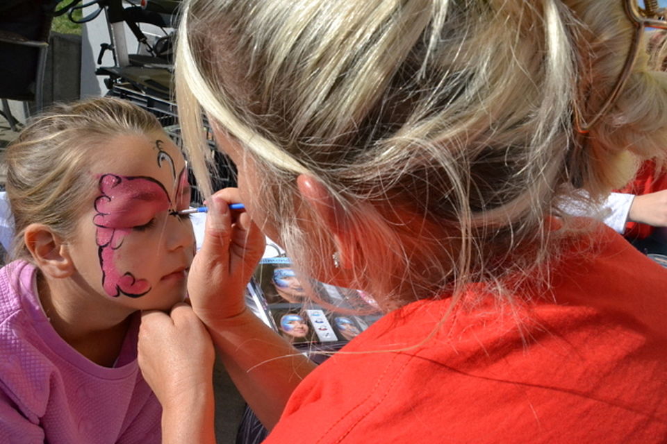 Bild zeigt ein kleines Mädchen (Larissa) welches an Schminktisch sitzt und von einer Kita-Mitarbeiterin einen pinkfarbenen Schmetterling ins Gesicht gemalt bekommt.