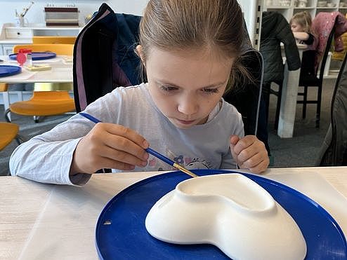 Bild zeigt ein Mädchen sitzend an einem Tisch. Vor ihr steht auf einer blauen Unterlage eine herzförmige Keramikschale. Sie hält einen Pinsel in der rechten Hand und malt auf der Schale.
