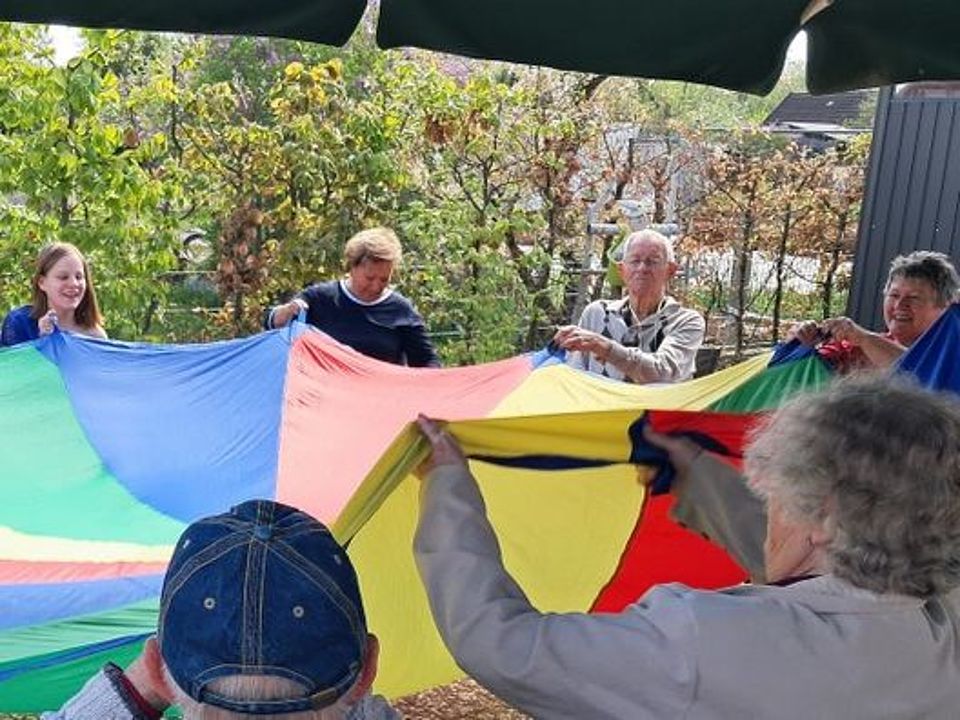 Bild zeigt mehrere Tagesgäste, die im Außenbereich auf der Terrasse mit einem Schwungtuch gemeinsam Bewegungs- und Koordinationsübungen machen. Es sind lachende und auch angestrengte Gesichter zu sehen.