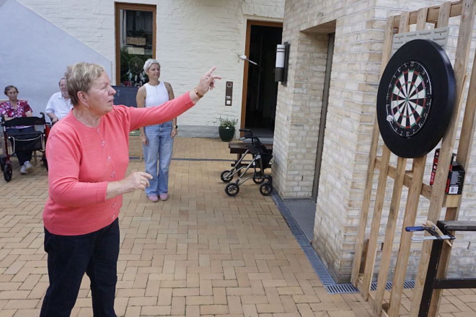 Bild zeigt einen weiblichen Tagesgast im Innenhof beim Darts spielen. Aus der linken Hand den Darts-Pfeil in Richtung Darts-Scheibe fliegen. Die Frau trägt einen roséfarbenen Pullover und eine schwarze Hose. Im Hintergrund sind weitere Personen zu sehen, welche gespannt dem Spiel folgen.
