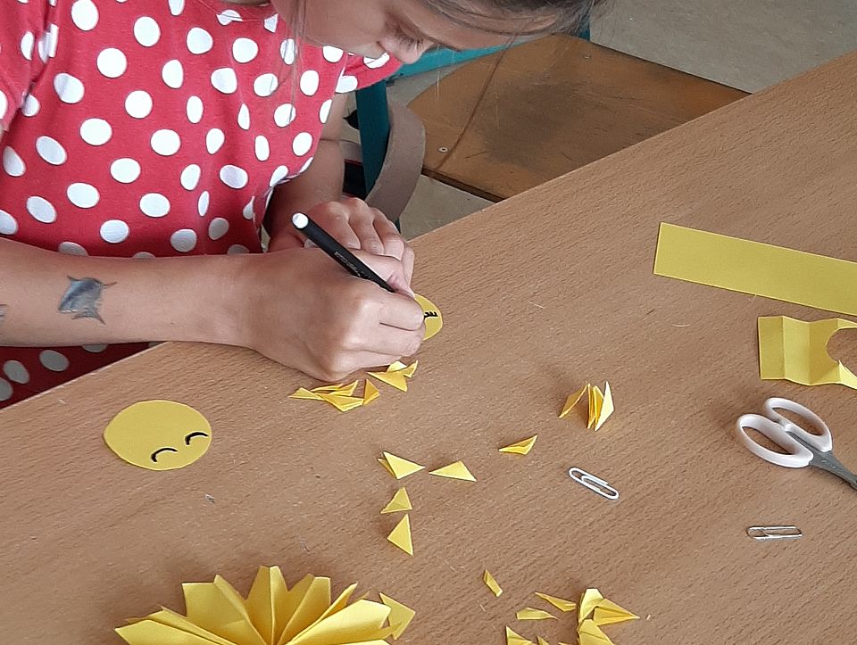 Bild zeigt ein Mädchen in einem rot-weiß karriertem Kleid. Sie sitzt an einem Tisch und bastelt mit gelbem Tonpapier. Auf dem Tisch liegen schon fertige Teile ausgeschnitten und eine Schere sowie das gelbe Tonpapier.