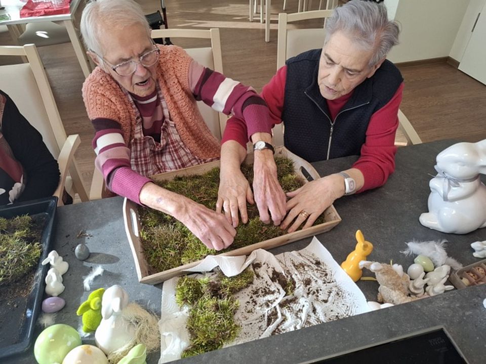 Bild zeigt zwei Frauen am Tisch sitzend. Vor ihnen steht eine Holzkiste mit Griffen an der Seite. Die Frauen legen Moos in die Kiste. Auf dem Tisch liegen und stehen verschiedene Osterdekoelemente wie Hasen und Kugeln und Eier.