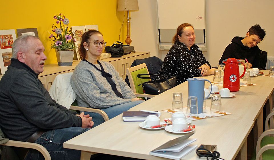 West Fest Planung Bild zeigt zeigt 4 Personen die an einem Tisch sitzen und über das West Fest reden. Auf dem Tisch stehen Kaffeekanne, Milch, Gläser und Tassen. Vor den Personen liegen Notizbücher und Schreiber. Im Raumhintergrund ist ein Sideboard zu sehen, auf dem ein Blumentopf und Bilder stehen.