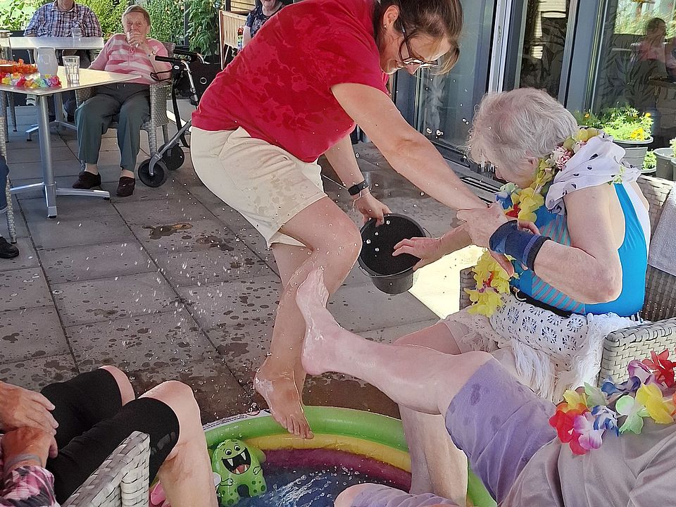 Bild zeigt: Eine Szene von einer sommerlichen Pool-Party im Freien. Eine jüngere Pflegekraft in einem roten DRK -T-Shirt steht lachend barfuß in einem kleinen, bunten, aufblasbaren Planschbecken und kippt Wasser aus einer schwarzen Schüssel über die Füße einer Seniorin, die auf einem Stuhl sitzt. Die Seniorin trägt eine blaue Badekleidung und eine Blumenkette, ebenso wie ein Herr, der rechts auf einem Stuhl sitzt und seine Füße im Wasser hat. Im Hintergrund sitzen weitere Personen entspannt an einem Tisch unter einer roten Markise, Sonnenschirm.