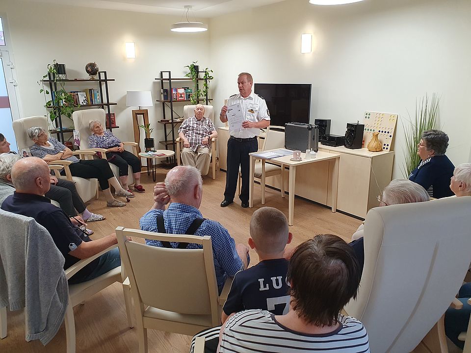 Bild zeigt: Ein Polizist in Uniform steht in der Mitte eines hellen Gemeinschaftsraumes und hält einen Vortrag zur Kriminalprävention. Er hält ein Informationsblatt in den Händen und spricht zu einer Gruppe von Senioren, die in hellen Sesseln in einem Halbkreis um ihn herum sitzen. Rechts von ihm steht ein kleiner Tisch mit einem Aktenkoffer. Im Hintergrund sind Regale mit Büchern und Pflanzen zu sehen.