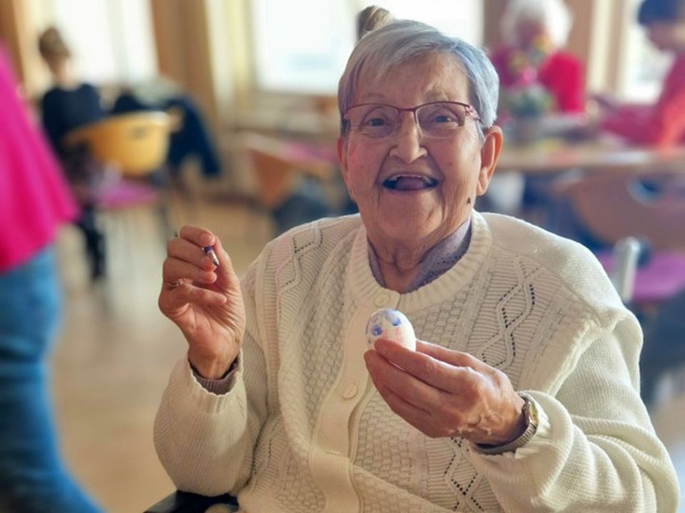 Bild 3 Bild zeigt eine sich freuende Seniorin, welche in der einen Hand ein fast fertig gemaltes Ei hält und in der anderen Hand einen kleinen Pinsel. Die Freude erkennt man an dem lachenden Gesicht. Im Hintergrund, leicht verschwommen, sind weitere Personen zu erkennen.