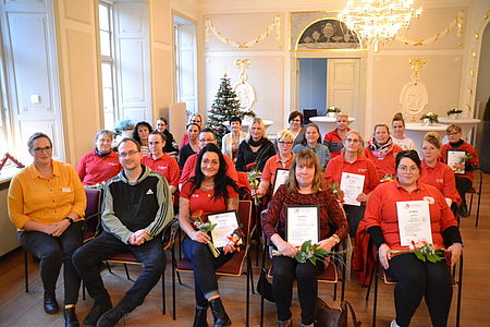 Weiterbildung Bild 1 Bild zeigt Mitarbeiter der Sozialstationen des DRK Kreisverband Güstrow e.V. im Saal des Palais 4+ in fünf Stuhlreihen sitzend. Etliche der Mitarbeitenden halten ihr Zertifikat der Ausbildung und einen Blumenstrauß in den Händen. Im Hintergrund ist der schon weihnachtlich geschmückte Saal zu sehen.
