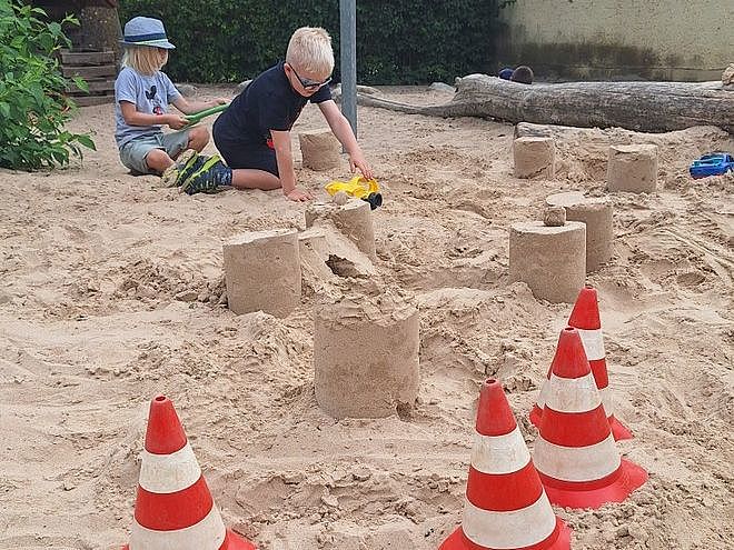 Bild zeigt: Zwei Jungen spielen in einem Sandkasten. Der vordere Junge in einem schwarzen T-Shirt und Sonnenbrille kniet und schiebt einen gelben Spielzeuglastwagen über eine im Sand gebaute Struktur. Hinter ihm, sitzend und einen Hut tragend, spielt ein anderer Junge mit einem grünen Spielzeug (Schaufel oder Rechen). Im Sand sind mehrere zylindrische Sandtürme oder -burgen gebaut. Im Vordergrund stehen drei rot-weiße Verkehrskegel (Pylonen). Im Hintergrund sind Büsche und ein großer Baumstamm zu sehen, was auf einen Spielplatz hindeutet.