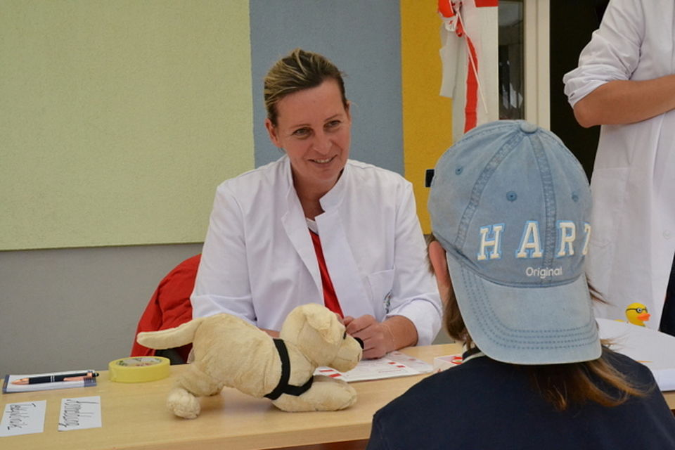 Bild zeigt einen Jungen (Aaron) mit rückwärtig aufgesetzter Schirmmütze von hinten vor dem Untersuchungstisch stehend. Am Tisch sitzt eine DRK-Mitarbeiterin in weißem Kittel und auf dem Tisch sitz der Plüschhund von Aaron, welcher gleich untersucht wird.