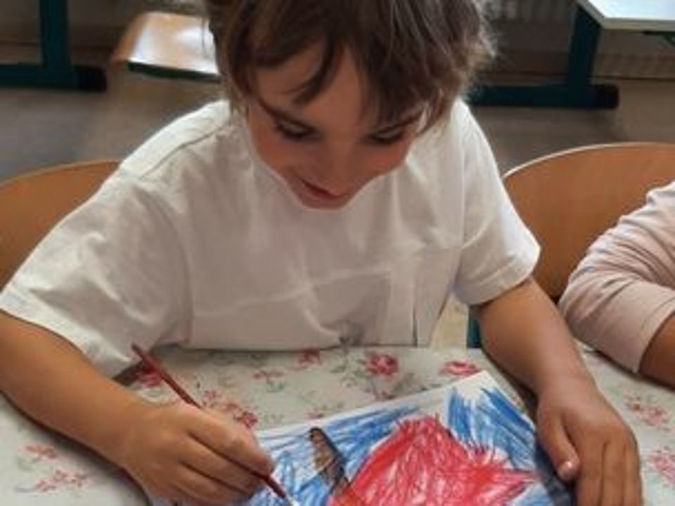 Bild zeigt einen Jungen an einem Tisch sitzend. Vor ihm liegt ein Blatt Papier auf dem mit viel Blau und Rot ein Bild gemalt ist. Vor dem Bild auf dem Tisch steht ein Kasten mit ganz vielen, farblich sortierten Malstiften
