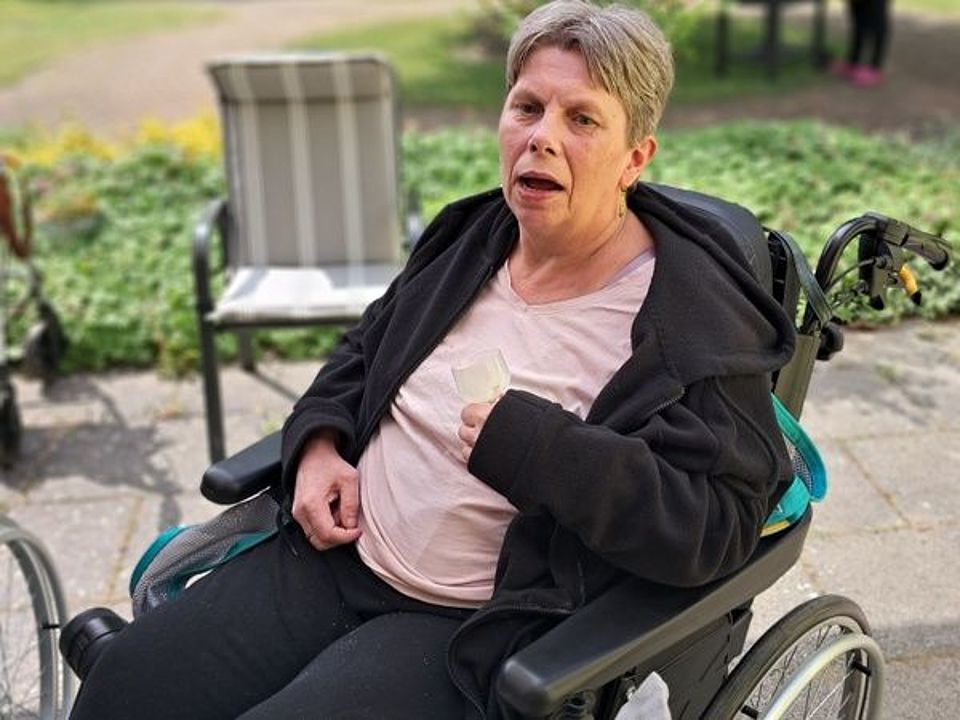 Bild zeigt eine Frau im Rollstuhl, welche auf der Terrasse steht, dass schöne Wetter und die frische Luft genießt.