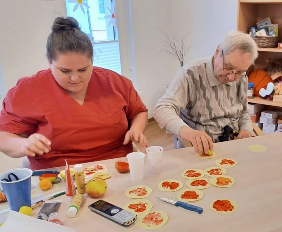 Bild zeigt eine Mitarbeiterin mit einem Tagesgast zusammen an einem Tisch sitzend und Papierteile mit einem Apfel bedruckend