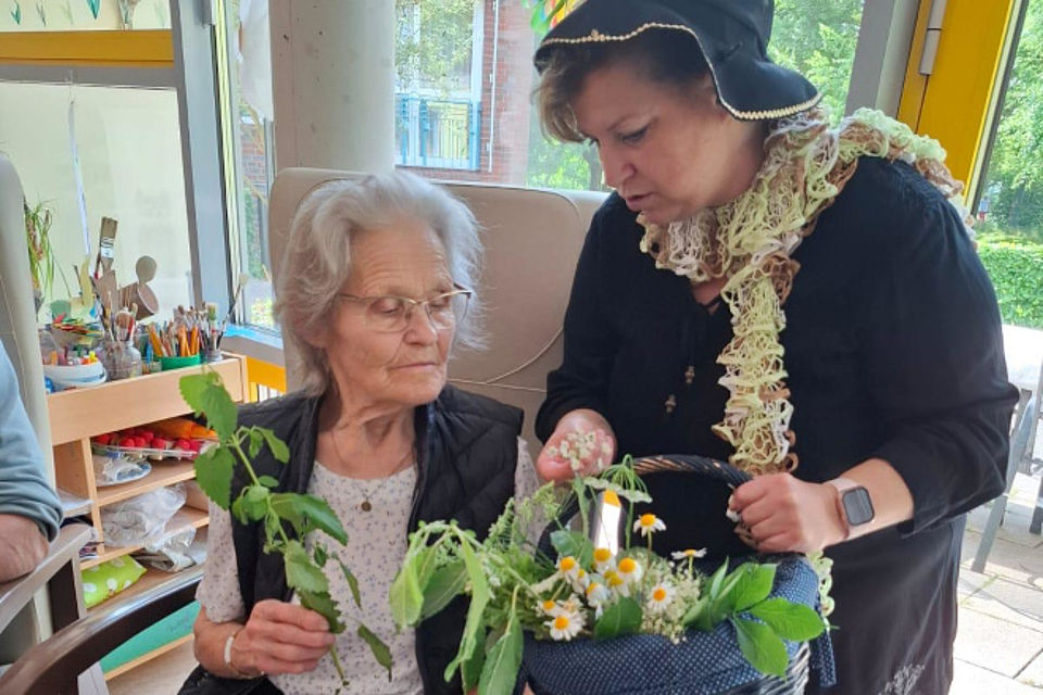 Bild zeigt: Eine Mitarbeiterin (rechts) in festlicher und thematischer Kleidung mit Hut und Schal zeigt einer älteren Dame (links) verschiedene Kräuter und Pflanzen aus einem Weidenkorb. Die Seniorin in der Mitte hält einen grünen Zweig in der Hand und betrachtet die Pflanzen und Kräuter konzentriert, um ihre Sinne anzuregen. Die Szene zeigt eine Aktivität zur Sinnesanregung im Innenraum der Tagespflege.