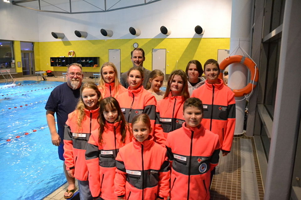 Juniorretter Bild 1 Bild zeigt eine Gruppe von 10 Kindern in DRK Softshelljacken mit dem Emblem des DRK und der DRK Wasserwacht, sowie dem Sponsoring-Patch der Güstrower Stadtwerke, stehend am Schwimmbeckenrand in der Güstrower Oase. Mit auf dem Bild ist links Hilmar Fischer, Ehrenamtskoordinator des DRK- Kreisverband Güstrow e.V. und im Hintergrund Herr Graßhoff Geschäftsführer der Güstrower Stadtwerke.