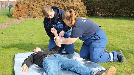 Bild zeigt ein Mädchen und einen Jungen der Jugendfeuerwehr Güstrow, bei der Versorgung eines "Verletzten", welcher auf einer Decke auf einer Wiese liegt. Die beiden Jugendfeuerwehrmitglieder verbinden den Arm des Verletzten.