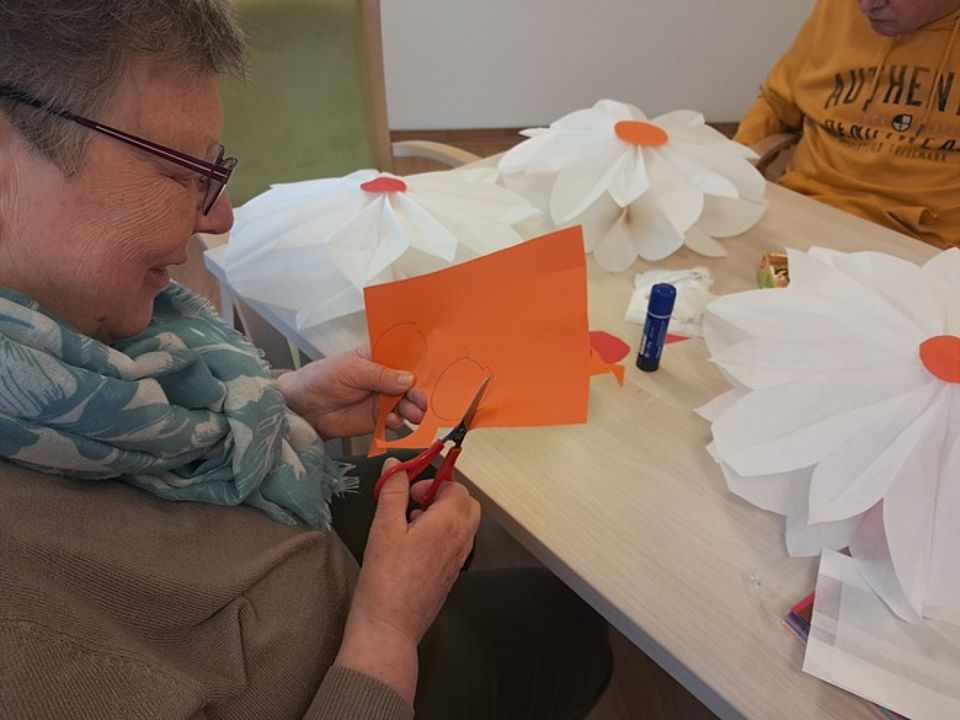 Bild zeigt eine Besucherin der Tagespflege beim ausschneiden von Kreisen aus einem orangefarbenen Stück Tonpapier. Diese Kreise kommen dann auf aus Frühstücksbeuteltüten gebastelte gr0ße Blumen