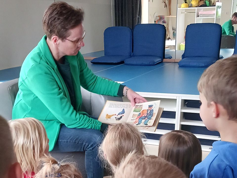 Vorlesewoche Bild 1 Bild zeigt Frau Henkel auf dem Boden sitzend mit einem Buch auf dem Schoß und liest aus dem Buch vor. Im Vordergrund sind Kinderköpfe von hinten zu sehen.