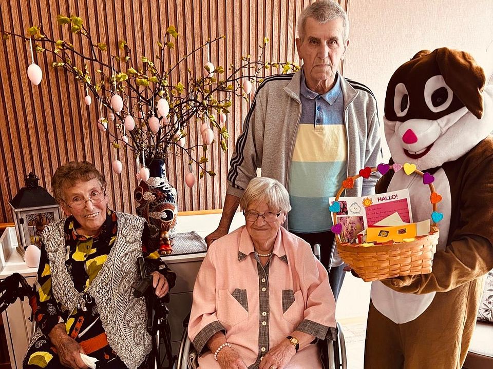 Bild zeigt 3 Bewohnende mit einem daneben stehenden "Osterhasen" (Mitarbeiter in einem Kostüm) vor einem bunt geschmückten Birkenstrauß. Der Osterhase hat ein Körbchen in den Pfoten, in dem die ganze Post liegt.