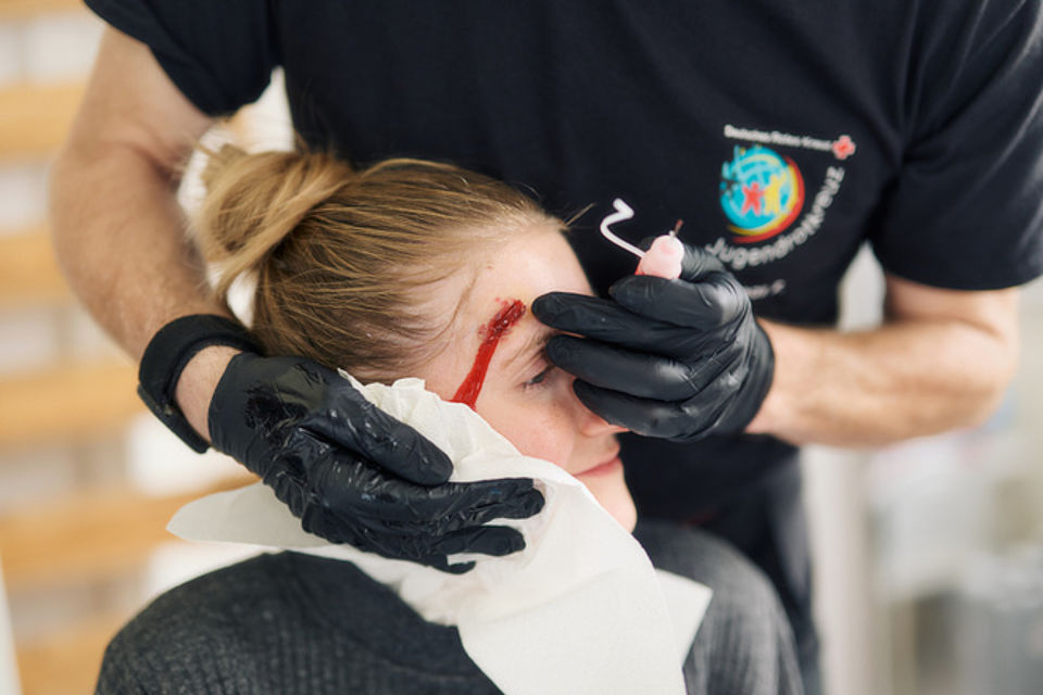 Notfalldarstellung Bild zeigt den Kopf einer jungen Frau, welche von einem Jugendrotkreuz Mitarbeiter eine Kopfplatzwunde über ihrer rechten Augenbraue geschminkt bekommt. Man sieht die recht realistisch aufgeschminkte Wunde, Kunstblut ist aufgetragen. Es läuft rechts am Auge vorbei Richtung rechtes Ohr. Der schminkende Mitarbeiter trägt schwarze Gummihandschuhe und hällt die Flasche mit dem Kunstblut in seiner linken Hand.
