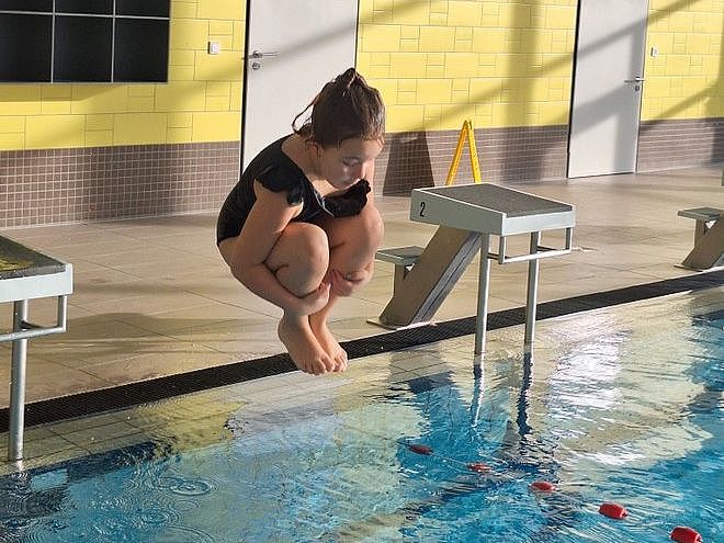 Bild zeigt ein kleines Mädchen, welches im Schwimmbad mit angehockten Beinen ins Wasser springt. Sie hat einen schwarzen Badeanzug an.