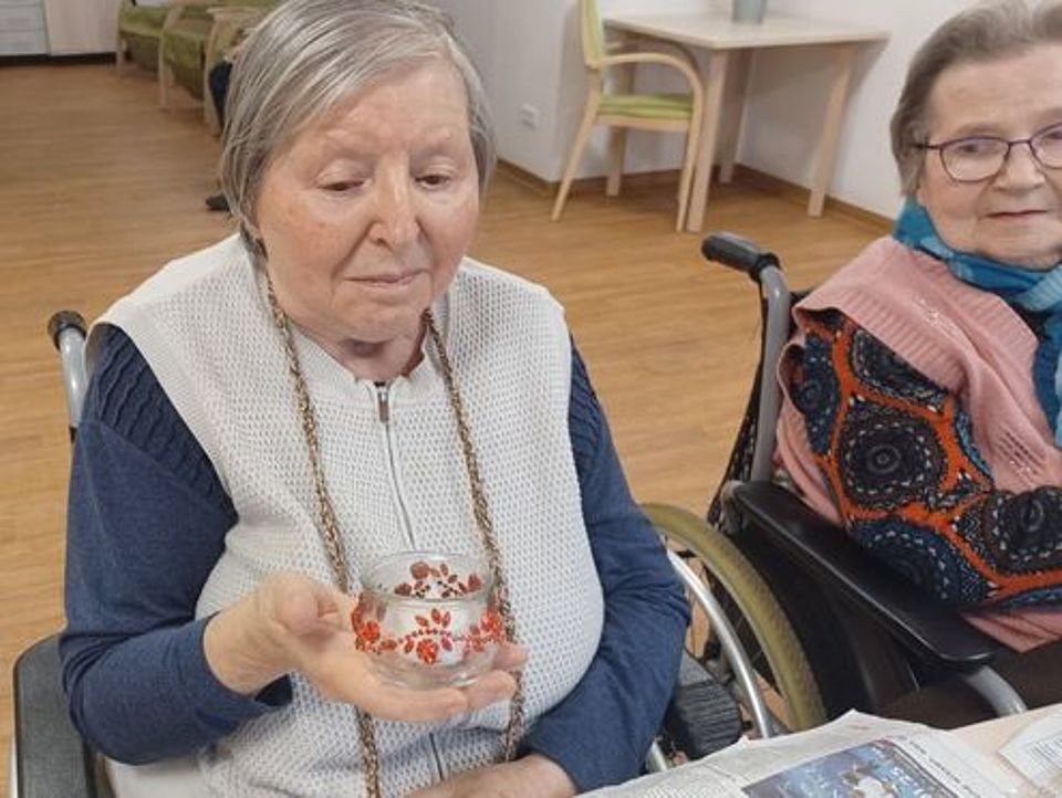 Bild zeigt Tagesgast Frau Marianne Hinniger am Tisch im Rollstuhl sitzend. In der rechten Hand hält sie das Windlicht, welches sie mit rotbraunen Blumenkettenornamenten verziert hat. Sie schaut etwas skeptisch auf ihr Werk.