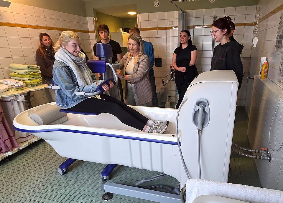 Bild zeigt eine Gruppe (8) von Menschen, die sich eine Pflegebadewanne mit Lifter anschauen. Eine junge Frau sitzt auf dem Lifter und testet wie sie damit in die Wanne kommt.