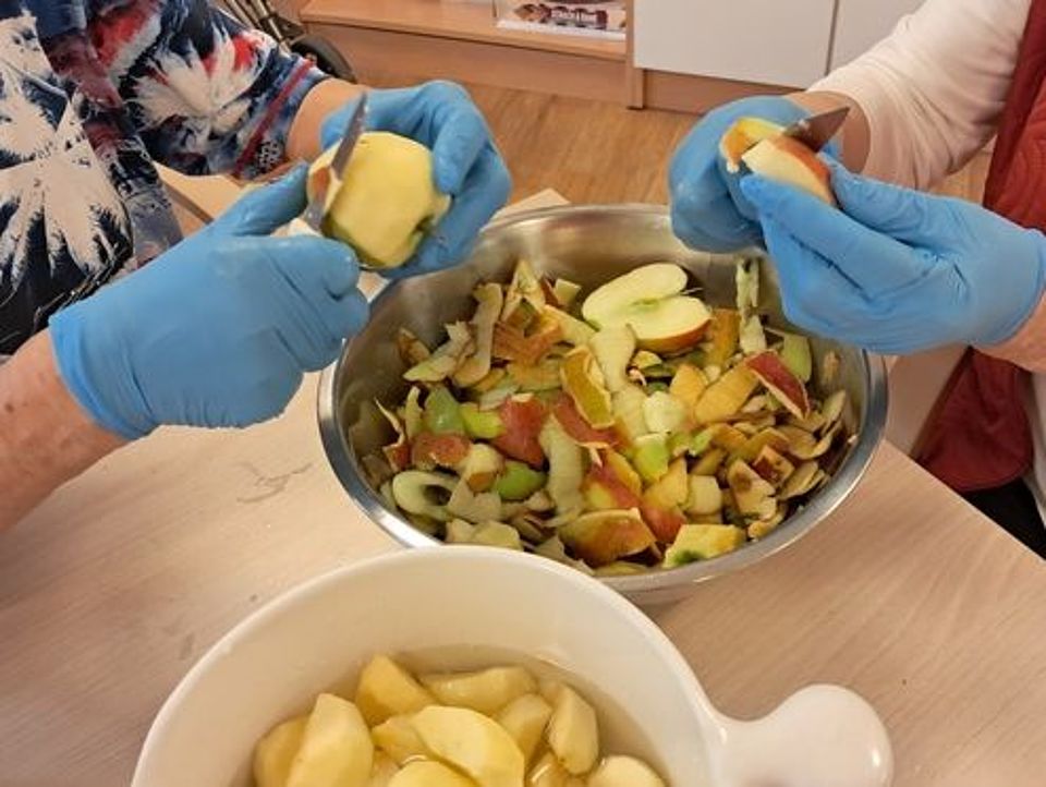 Bild zeigt eine Edelstahlschüssel auf einer Tischecke über der zwei Handpaare Äpfel schälen. Im Vordergrund ist eine weiße Keramikschüssel halb zu sehen, in der die geschälten Äpfel liegen.