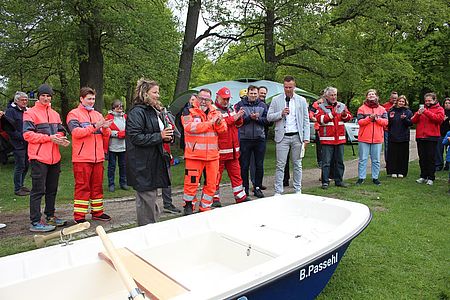 Bild zeigt die Taufe des neuen Wasserwacht Bootes für den Inselsee. Es stehen viele Menschen um das Boot und applaudieren zur Taufe.