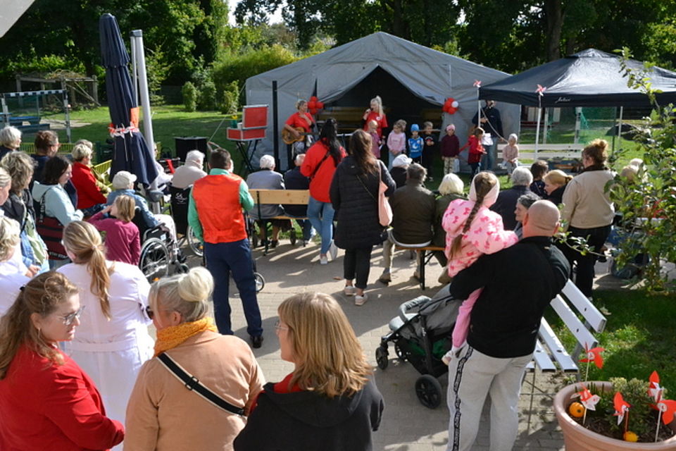 Bild zeigt den Außenbereich der Kita mit der Terrasse. Im Hintergrund steht ein großes Festzelt vor dem eine Gruppe von Kita-Kindern mit zwei Erzieherinnen stehen und ihr Programm vortragen. Viele Menschen sind auf der Terrasse und schauen den Kindern zu.