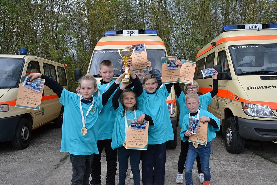 Bild zeigt die JRK Gruppe Altersstufe 0 mit Pokal Urkunden und Medaillen. Sieben Kinder stehen vor Rot-Kreuz-Fahtzeugen und halten stolz und fröhlich ihre Urkunden und den Pokal in die Kamera
