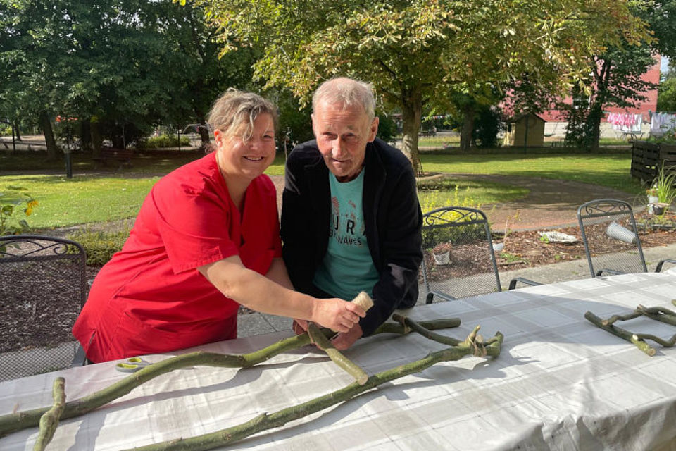 Bild zeigt: Eine Mitarbeiterin in roter Dienstkleidung und ein älterer Mann bauen gemeinsam Dekoration im Freien auf einem Gartentisch. Beide konzentrieren sich darauf, Äste mit einem Stück Seil oder Band zusammenzubinden, um ein Dekorationsobjekt zu erstellen. Der Mann trägt ein hellblaues Shirt und eine dunkle Jacke. Die Szene zeigt eine gemeinsame kreative Aktivität und Bastelarbeit im Grünen.