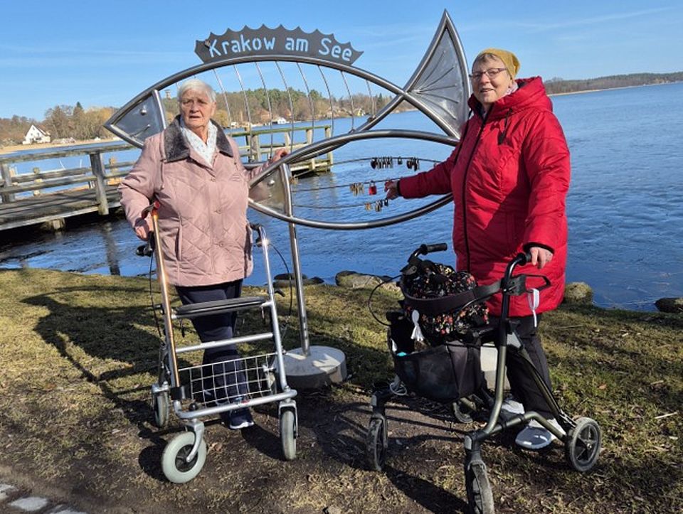 Bild zeigt zwei Tagesgäste beim Spaziergang an der Seepromenade. Sie stehen mit ihren Rollatoren vor einem aus Edelstahl gefertigten großen Fisch, der in seiner Rückenflosse Krakow am See stehen hat. Im Hintergrund ist der Krakower See zu sehen.