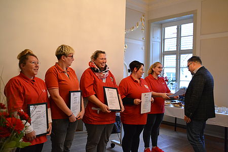 Bild zeigt 5 Frauen, die ihr Abschlußzertifikat überreicht bekommen. Alle tragen ein rotes DRK Shirt. 4 Frauen halten die Urkunde vor sich. die 5. Frau erhält gerade ihre Urkunde aus den Händen des Vorstandsvorsitzenden  Alexander Plass überreicht.