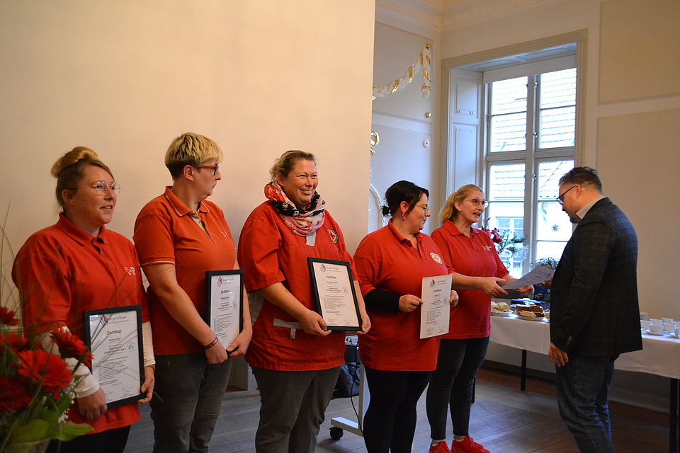 Quereinstig Bild 1 Bild zeigt 5 Frauen, die ihr Abschlußzertifikat überreicht bekommen. Alle tragen ein rotes DRK Shirt. 4 Frauen halten die Urkunde vor sich. die 5. Frau erhält gerade ihre Urkunde aus den Händen des Vorstandsvorsitzenden Alexander Plass überreicht.