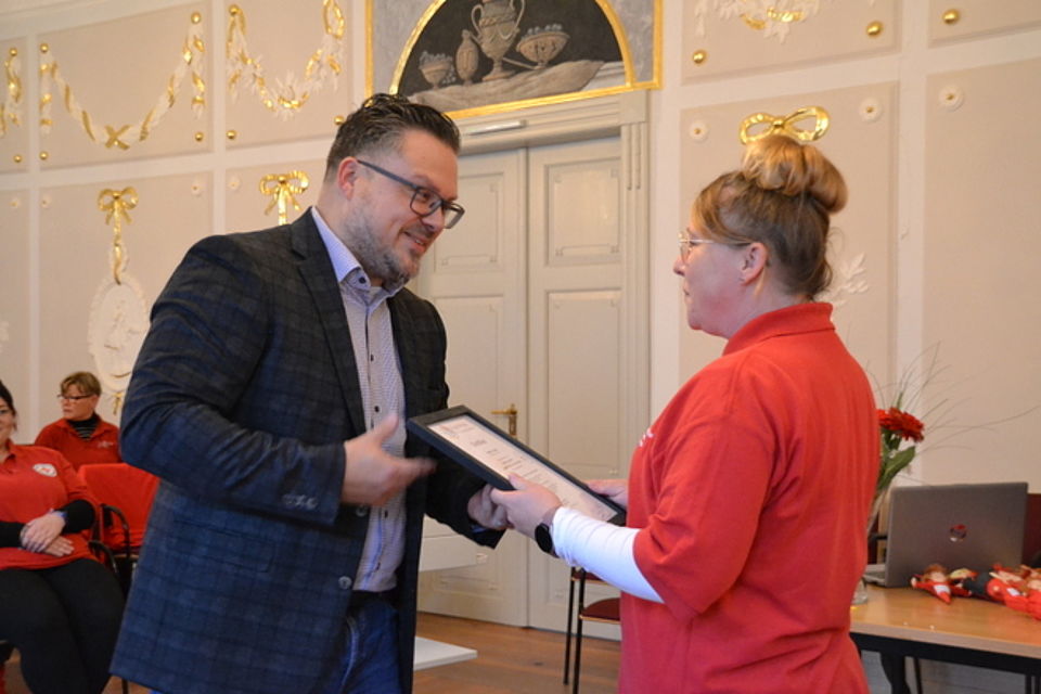 Weiterbildung Bild 3 Bild zeigt Alexander Plass, Vorstand des DRK-Kreisverband Güstrow e.V., bei der Übergabe der Zertifikatsurkunde an eine Mitarbeiterin. Sie trägt ein rotes DRK-Shirt und ist sichtlich gerührt.