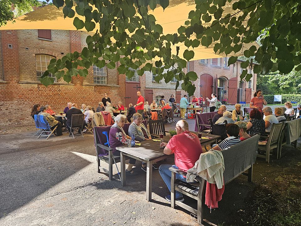 Bild zeigt: Ein sommerliches Grillfest auf dem Gutshof Hohen Luckow. Zahlreiche Tagesgäste und ihre Angehörigen sitzen im Freien an Holztischen und Bänken. Sie sind durch ein großes, beigefarbenes Sonnensegel und natürliche Baumkronen vor der Sonne geschützt. Im Hintergrund ist ein rustikales Backsteingebäude mit großen Torbögen und Fenstern zu sehen, wo sich der Grill- und Buffetbereich befindet.