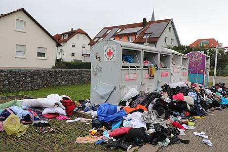 Bild zeigt 5 DRK Kleidercontainer auf einer Stellfläche. Davor liegen Unmengen an Kleidungsstücken. Der Haufen dieser Kleidungsstücke zieht sich über die gesamte Breite aller Kleidercontainer und ist und ist einen guten Meter hoch.