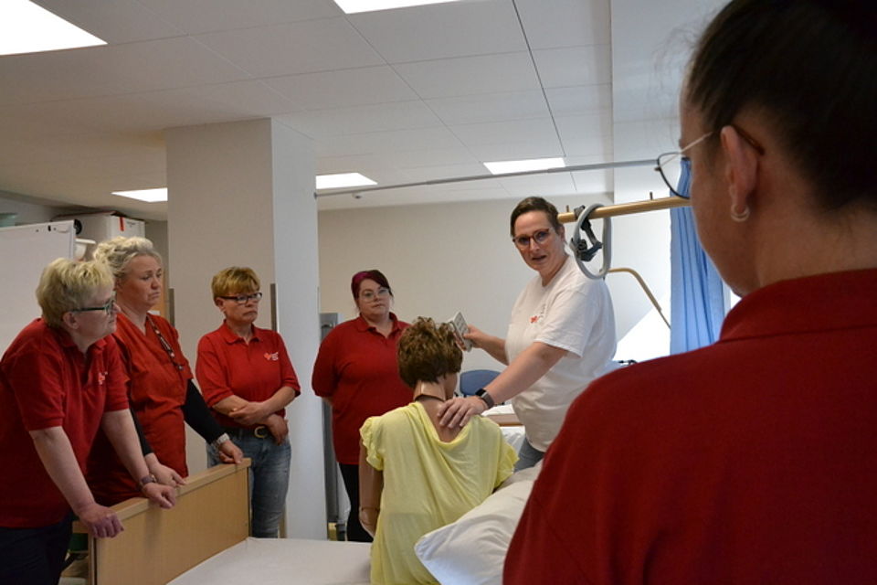 Bild zeigt eine Gruppe von auszubildenden Pflegefachkräften, vor einem Pflegebett im Halbkreis stehend. Frau Behlke, Ausbilderin, erklärt und zeigt bestimmte Handgriffe, wie mit bettlägerigen Patienten umzugehen ist.