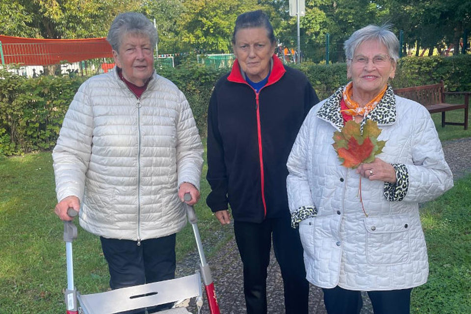 Bild zeigt: Drei ältere Damen stehen nebeneinander im Freien vor einem grünen Buschwerk und Bäumen. Die Frau ganz links stützt sich auf einen Rollator und trägt eine hellbeigefarbene Steppjacke. Die Dame in der Mitte trägt eine dunkle Fleecejacke. Die Frau rechts hält ein buntes, rotes und gelbes Herbstblatt in der Hand und trägt eine weiße Steppjacke mit Leopardenmuster an Kragen und Ärmeln. Die Szene vermittelt einen gemeinsamen Herbstspaziergang oder Ausflug der Seniorinnen.