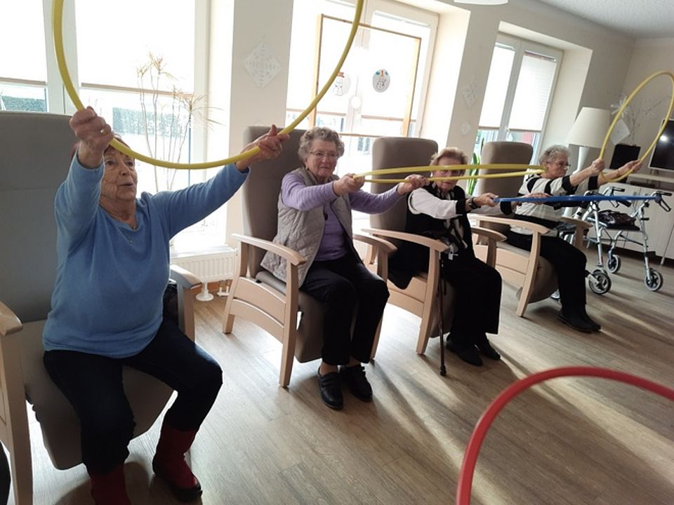 Bild zeigt 4 Besucherinnen der Tagespflege in den geräumigen Sesseln sitzend und mit Sportringen leichte gymnastische Übungen machen. Die Ringe werden mit beiden Händen vor dem Oberkörper hoch und runter bewegt.