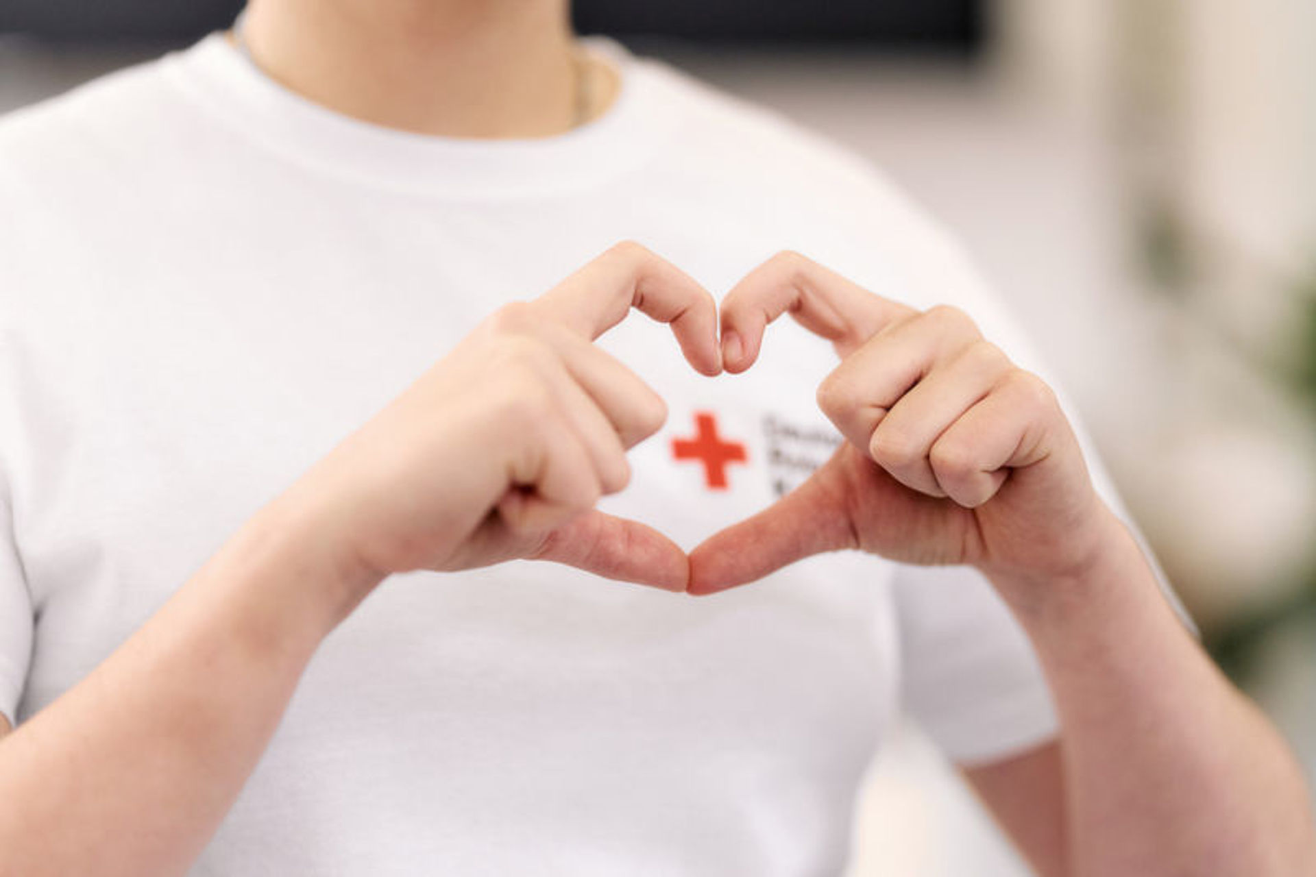 Bild zeigt nur den Oberkörper eines jungen Mannes mit einem weißen T-Shirt mit DRK Logo, welcher die Finger beider Hände zu einem Herz geformt vor das DRK Logo hält