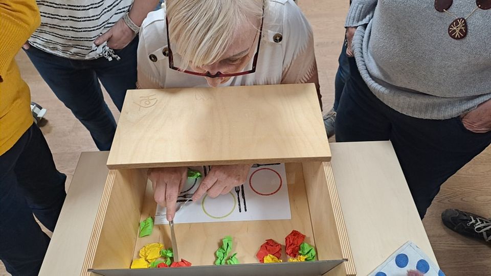 Bild zeigt wie Teilnehmende mit einer Sinnenwahrnehmungsbox versuchen Essen auf einen Teller zu füllen.