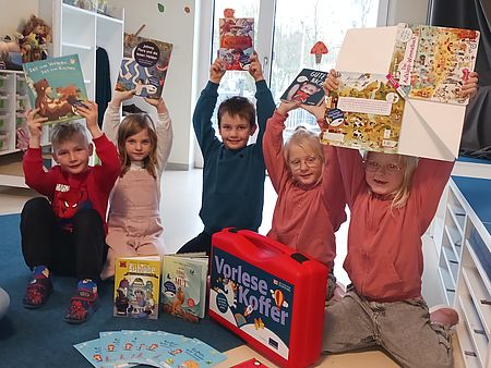 Bild zeigt 5 Kinder (3 Mädchen 2 Jungen), welche jedes ein Buch in den Händen über ihren Kopf in die Höhe streckt. Vor den Kindern, die auf dem Boden sitzen, steht ein Vorlesekoffer, dessen Inhalt (Bücher, Broschüren und interaktives Material) vor ihnen ausgebreitet ist. Auch die Bücher in den Händen der Kinder kommen aus diesem Koffer.
