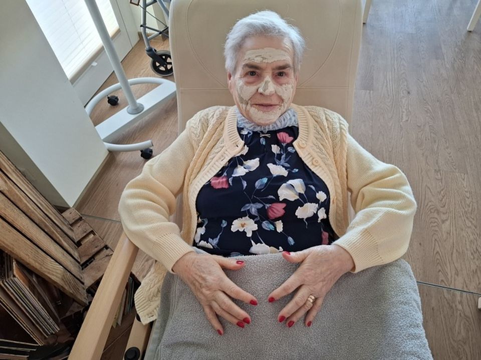 Bild zeigt eine Frau auf einem Ruhesessel liegend. Sie hat im Gesicht eine Maskencreme aufgetragen. Sie hat graue Haare und trägt eine dunkle Bluse mit Blumenmotiven. Darüber eine Strickjacke in einem beige-pfirsich Ton. Bis zur Hüfte ist sie mit einer grauen Decke zugedeckt. Darauf liegen ihre Hände mit frisch lackierten roten Fingernägeln.