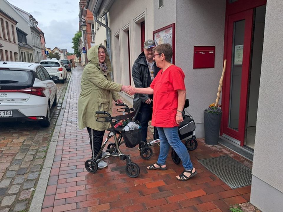 Bild zeigt eine Besucherin der Tagespflege vor dem Haus wo sie von einer Mitarbeiterin mit Handschlag begrüßt wird. Leider regnet es ein wenig.