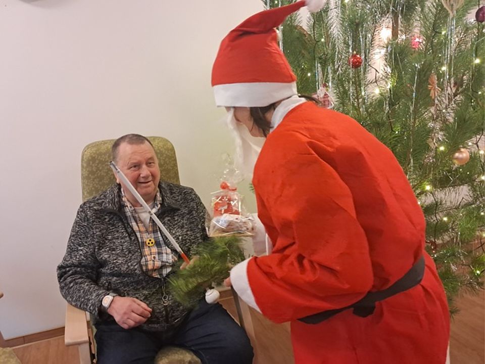 Bild zeigt eine Mitarbeiterin als wEIHNACHTSMANN verkleidet wie sie einem Tagesgast ein Geschenk überreicht. Rechts im Hintergrund ist ein geschw2ückter Weihnachtsbaum zu sehen.