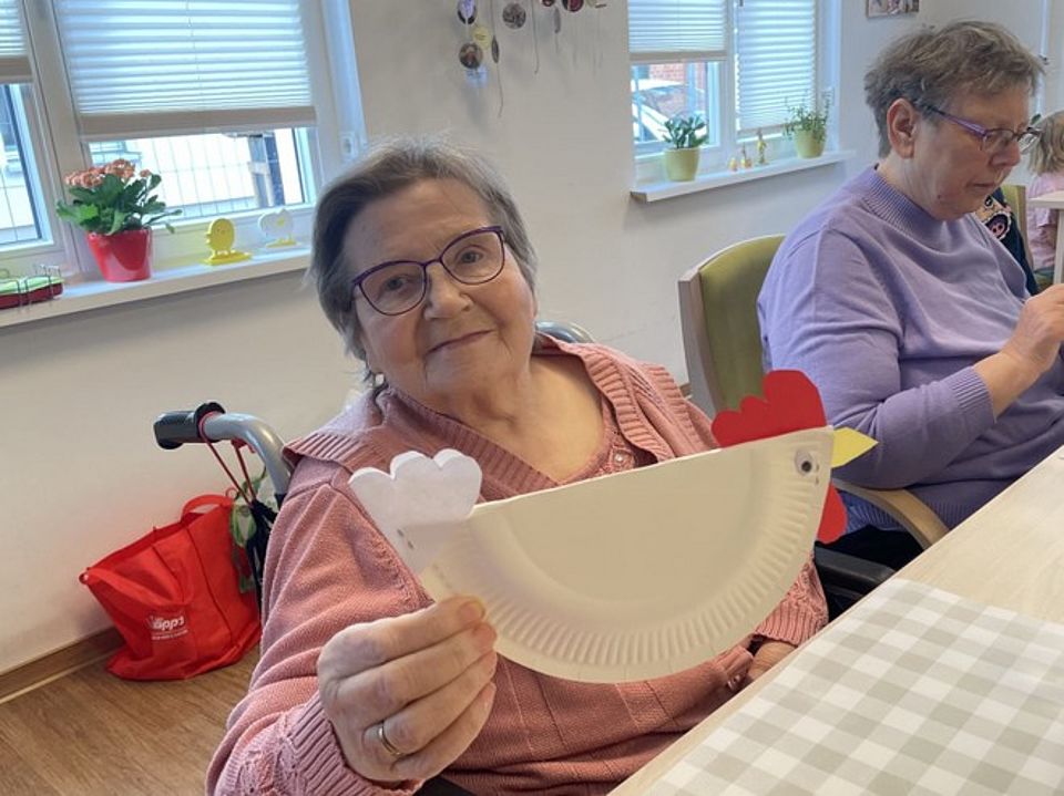 Bild zeigt zwei Besucherinnen der Tagespflege an einem Tisch sitzend und aus Papptellern Osterdekoration basteln. Hier ist es ein Huhn geworden