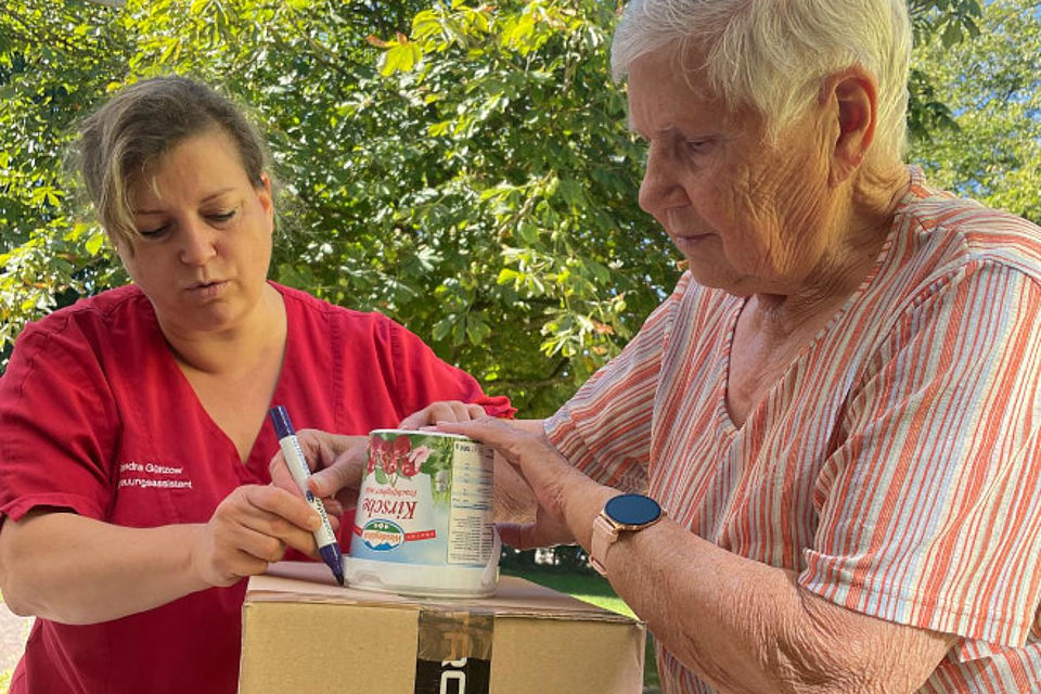 Bild zeigt: Eine Mitarbeiterin der Tagespflege in roter Arbeitskleidung (links) und eine ältere Dame (rechts) gestalten einen Karton im Freien. Die Mitarbeiterin hält einen Filzstift in der Hand und schreibt auf einen Karton, auf dem ein leerer großer Becher mit Kirschjoghurt steht. Die ältere Dame in einem gestreiften Oberteil und mit einer Smartwatch beobachtet und unterstützt durch festhalten des Bechers.